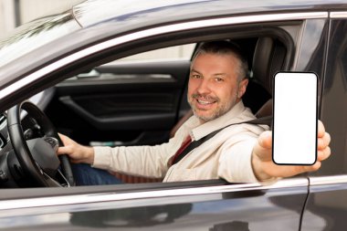 Gülümseyen orta yaşlı adam arabada oturuyor, beyaz ekranlı akıllı telefonu gösteriyor, seyrüsefer için mobil uygulama gösteriyor, modelleme yapıyor.