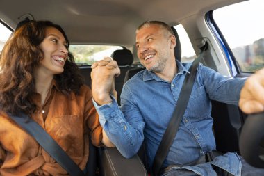 Avrupalı çift ön koltuklarda oturup arabalarında kemerlerini bağlıyor, el ele tutuşuyor ve birbirlerine gülümsüyor. Turizm, gezi, yolculuk