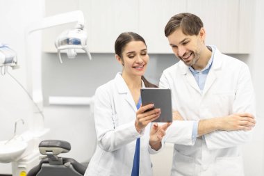 Modern diş kliniğinde dijital tablet ekranına bakan profesyonel erkek doktor ve kadın asistan, hasta tedavisini tartışıyor