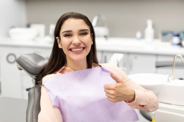 Profesyonel diş tedavisi gördükten sonra baş parmağını gösteren mutlu genç bayan hasta modern diş kliniğinde sandalyede oturuyor.
