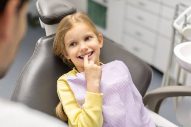 Pediatrik dişçilik kliniğindeki randevu sırasında dişçi koltuğunda oturan ve dişlerini işaret eden küçük bir kız hasta.