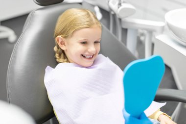 Güzel küçük Avrupalı kız aynaya bakıyor ve dişçiye gittikten sonra gülümsüyor, check-up yaptırıyor ya da diş cilası yaptırıyor.