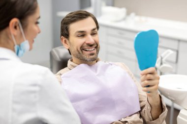 Diş hekimliği muayenesi sırasında diş tedavisinin sonuçlarından memnun olan mutlu adam diş aynasına bakıyor.