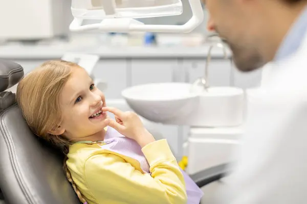 Bebek dişlerini erkek pediatri doktoruna gösteren, gülümsemesini işaret eden, klinikteki koltukta oturan sevimli küçük kız.