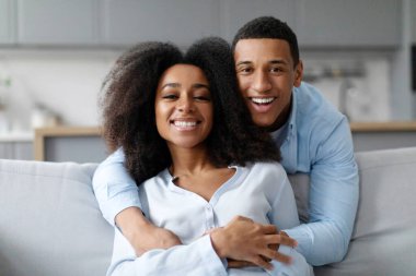 Mutlu siyah adam karısını kucaklıyor ve kameraya gülümsüyor, aile kanepeye oturuyor, hafta sonunun tadını çıkarıyor. Evlilikte mutluluk.