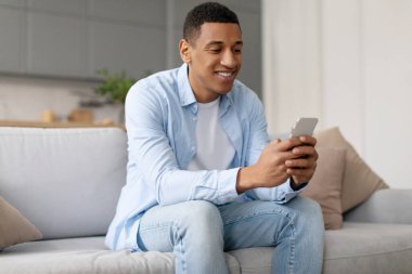 Akıllı telefon kullanan, internete takılan, mesajlaşan, evdeki koltukta oturan gülümseyen siyahi adam. Yeni havalı uygulama kullanan adam