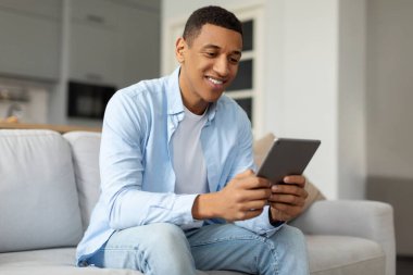 Dijital tabletle koltukta oturan mutlu Afrikalı Amerikalı adam, evdeki modern bloknotlarda flört uygulaması kullanıyor.