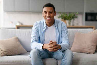 Evdeki kanepede oturan ve kameraya gülümseyen, iç mekan manzaralı Afro-Amerikan genç adamın portresi.