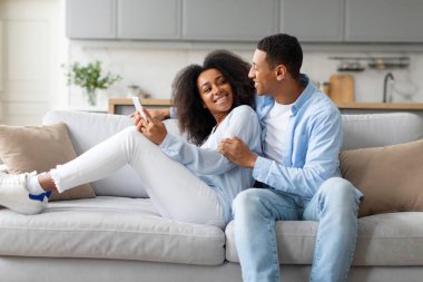 Mutlu siyah çift akıllı telefon kullanıyor, evdeki koltukta dinleniyor, yeni mobil uygulama kullanıyor, internette geziniyor, sosyal medyayı kontrol ediyor.