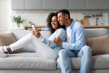 Genç siyahi çift sosyal medyayı cep telefonlarıyla paylaşıyor, koltukta oturuyor, alet kullanıyor, akıllı telefonlara bakan kadını kucaklıyor.