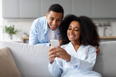 Neşeli siyahi adam kadının arkasında duruyor ve cep telefonuna bakıyor. İçeriği paylaşan kadın, koltukta oturuyor.