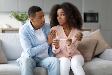 Genç siyah koca depresyondaki karısını negatif hamilelik testiyle rahatlatıyor, evdeki kanepede oturuyor. Kısırlıktan muzdarip eşler