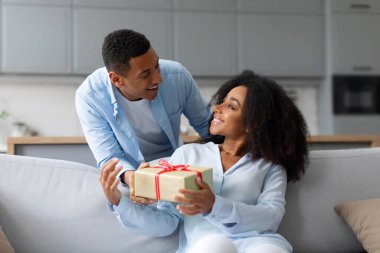 Arkasından sarılan ve karısına hediye kutusu veren Afrikalı Amerikalı bir adam. Doğum günü için büyük bir sürpriz hazırlıyor. İç mekan, serbest alan.