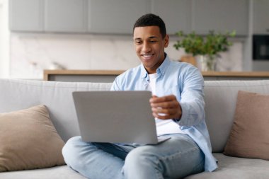 Serbest yaşam tarzı ve çevrimiçi uzaktan çalışma konsepti. Mutlu Afrikalı Amerikalı adam dizüstü bilgisayar kullanıyor, uzaktan çalışıyor, evdeki kanepede oturuyor..