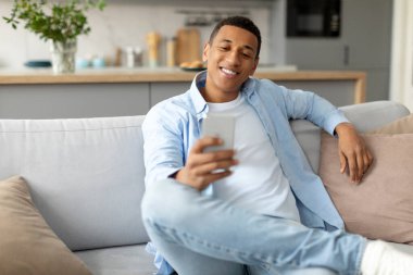 Gülümseyen siyah adam cep telefonu tutuyor, SMS mesajı yazıyor ya da internette sörf yapıyor, oturma odasında kanepede oturuyor, boşluğu kopyalıyor.