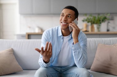 Mutlu siyah adam cep telefonuyla konuşuyor ve gülümsüyor, evdeki koltukta oturuyor, telefonla iletişim kuruyor, hoş sohbetler ediyor.