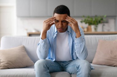 Stresli genç siyah adam evdeki kanepede oturuyor, tapınakları ovuyor, baş ağrısı çekiyor ya da düşünüyor.
