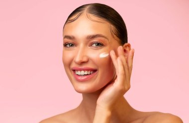 Gülümseyen şehvetli esmer kadın genç parlak cilt için yüz kremi kullanıyor, pembe stüdyo arka planında izole edilmiş poz veriyor.