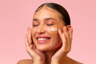 Yüz derisi. Güzel mutlu bayan yüzüne nemlendirici sürüyor ve kapalı gözlerle gülümsüyor, pembe arka planda kozmetiğin tadını çıkarıyor.