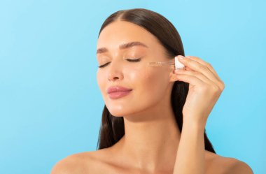 Güzel genç bir bayan nemlendirici yüz serumu uyguluyor. Günlük güzellik prosedürleri uyguluyor. Düşük yağlı, mavi arka planlı.