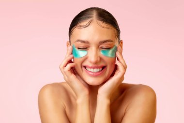 Yeşil göz bantlı neşeli genç bir kadın. Gözleri kapalı gülüyor, pembe arka planda poz veriyor. Sabah güzellik rutini, kozmetik prosedür