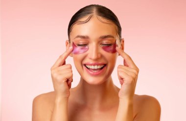 Gözlerinin altına pembe bir yama takmış ve gülümseyen güzel bir genç kadın, pembe arka planda yalnız poz veriyor. Cilt bakımı, kozmetik prosedürleri konsepti