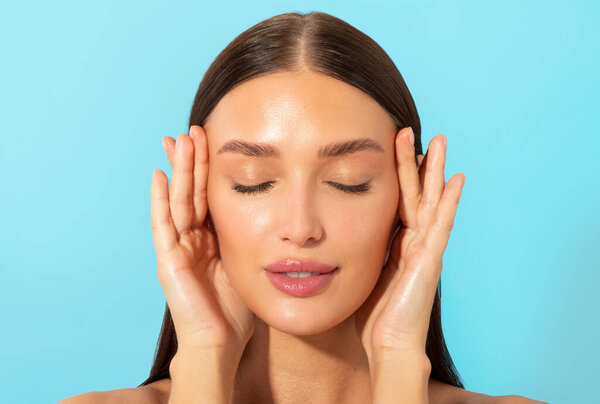 Close-up portrait of attractive European woman touching temples, showing her perfect glowing clean skin, posing isolated over blue background