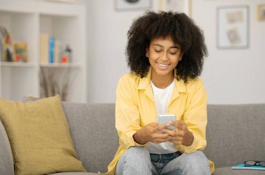 Evdeki koltukta otururken cep telefonu kullanan mutlu, genç siyahi bayan, en yeni flört uygulamasında sohbet ediyor, sosyal medyayı kontrol ediyor, kopyalama alanını kopyalıyor.