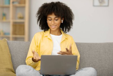 Laptop bilgisayarından arayan, kulaklık takan, webcam 'e konuşan ve el kol hareketi yapan, evdeki koltukta oturan genç siyahi kadın videosu. Video çağrısı, sanal sohbet kavramı