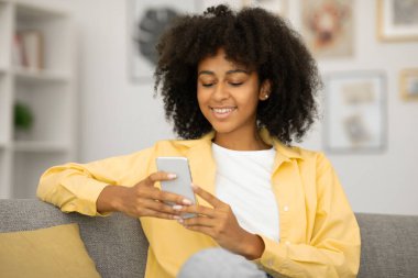 Genç siyahi kadın akıllı telefondan mesaj atıyor, mesaj okuyor ve oturma odasında gülümsüyor. Gadget ve sosyal ağlar, başarılı günlük