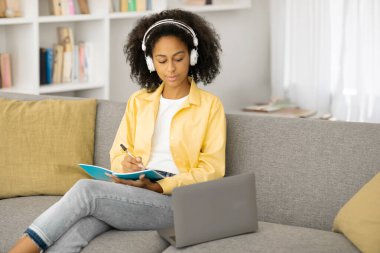 Kulaklık takıp bilgisayar kullanarak kanepede oturan ve notlar alan, online derslere katılan kız öğrenci.