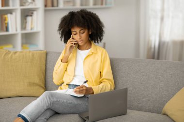 Serbest meslek kavramı. Siyahi bir kadın cep telefonuyla konuşuyor, not alıyor, kanepede dizüstü bilgisayarla oturuyor. Kadın arama işi