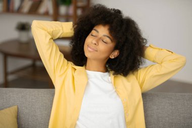 Sakin, siyah bir kadın arkasını yaslıyor, ellerini başının arkasına koyup evdeki rahat kanepeye oturuyor.