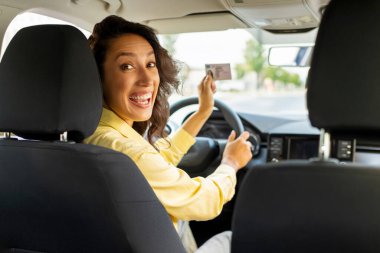 Çok mutlu bir kadın kendi arabasında oturuyor ve ehliyetini gösteriyor, sınavı geçişini kutluyor, sürücü kursunu bitiriyor, aracının içinde oturuyor.
