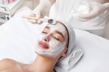 Güzellik uzmanının eli kadın yüzüne kozmetik maske takıyor, bayan müşteri güzellik salonunda cilt bakımı yaptırıyor. Cilt bakımı ve yüz bakımı kavramı