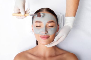 Rahatlamış genç bir kadın spa salonunda profesyonel güzellik uzmanından cilt bakımı için yüz soyma maskesi alıyor.