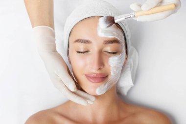 Kozmetik uzmanı, masadaki güzel bayanın yüzüne beyaz kilden maske takıyor güzellik salonundaki güzellik tedavisinden zevk alıyor.