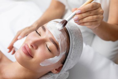 Spa uzmanı fırça tutuyor ve spa salonunda uzanan ve cilt bakımını seven güzel genç bayana beyaz besleyici maske yapıyor.