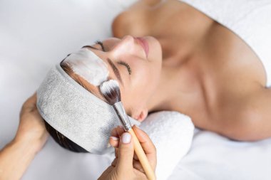 Güzellik salonunda güzellik tedavisinden hoşlanan genç bir bayan yüzüne kilden bir maske takıyor.