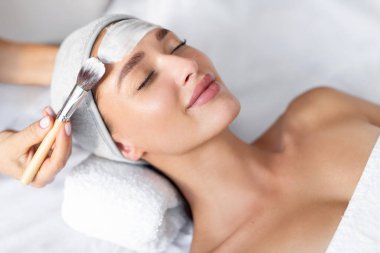 Güzeller güzeli genç bir kadının güzellik salonunda uzanırken maske takması ve cilt bakımı prosedürlerinin keyfini çıkarması.