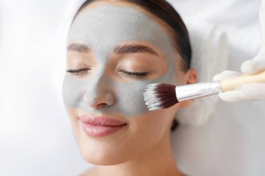 Kozmetik uzmanı, genç kadınların yüzüne gri kilden maske takıyor, bayan müşteri, kapalı gözlerle dinleniyor, yakın plan.