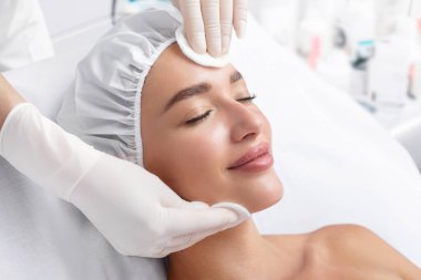 Kozmetikçiyle randevusu sırasında yüzü pamuk pedlerle temizlenirken gözleri kapalı yatıp gülümseyen huzurlu ve rahat genç bir bayan.