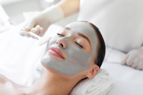 Woman getting professional facial care by beautician at spa salon, lady lying with grey clay mask. Skincare procedure