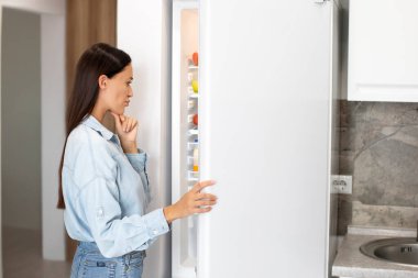 Düşünceli genç bir kadın buzdolabını açıp ne yiyeceğini düşünüyor, içine bakıyor ve ciddi bir ifadeyle çenesine dokunuyor.