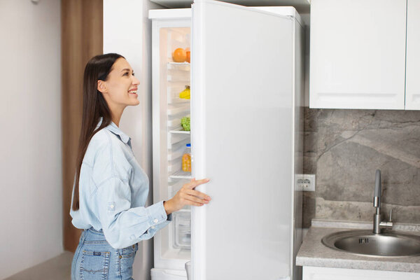 Lady looking inside of fridge while standing in kitchen, opening refrigerator and searching for food, rear view with copy space