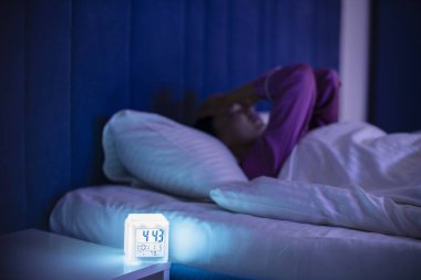 Uykusuzluk çeken stresli bir kadın, geceleri yatakta uyanık ve elleriyle yüzünü örtüyor, dijital alarm lambasına odaklanıyor.