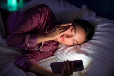 Akıllı telefondan video izlerken gözlerini ovuşturan genç bir kadın gecenin geç saatlerinde karanlık yatak odasının içinde uzanıyor.