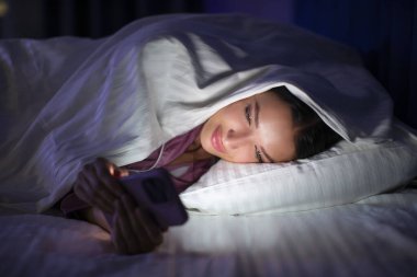 Geceleri romantik sohbetler. Cep telefonunda cep telefonu uygulaması kullanan mutlu bir kadın karanlık yatak odasındaki battaniyenin altında yatıyor.