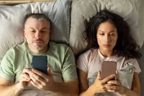 Stock image Spouses ignoring each other, lying on bed and using their phones, top view. Treason, addiction on social networks and technology