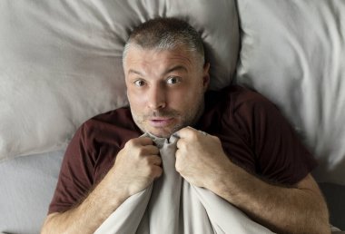 Korkmuş orta yaşlı bir adam battaniye tutuyor ve kameraya bakıyor, yatak odasında, manzaranın üstünde yatıyor. Panik atak, korku ve endişe, zihinsel sağlık sorunları kavramı